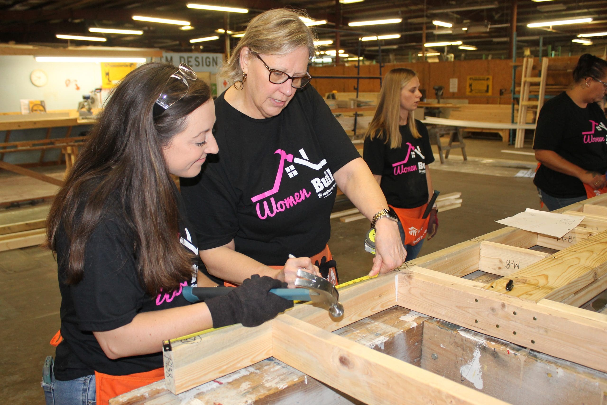 Two women volunteers in pink hard hats and Women Build shirts hammering together